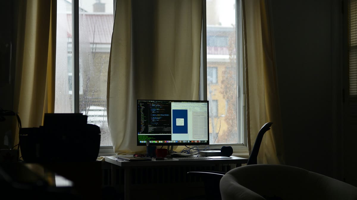 Stůl u okna s monitorem, na kterém je kód a náhled rozvržení; venku obyčejná ulice se žlutým domem za šedivého dne.